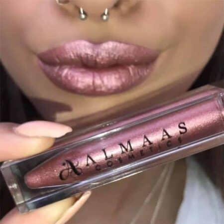 תקריב של שפתיים ורודות מטאליות עם פירסינג באף, כשהן אוחזות בשפתון מטאלי עמיד בגוון 65 של ALMAAS Cosmetics בגוון ורוד מנצנץ תואם, המונח ליד הפה. | אלמאס קוסמטיקה – מתנה מושלמת של טיפוח ויוקרה