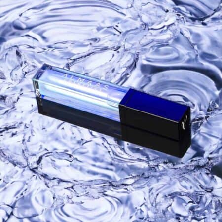 שפופרת גלוס לשפתיים מלבנית בצבעי כחול ושקוף, שעליה הכיתוב "מנפח שפתיים פנינה", מונחת באלכסון על פני מים נוצצים ומרצדים; מכסהה הכהה יוצר ניגוד עם הרקע המנצנץ ויוצר השתקפויות המוסיפות אפקט מבריק ומרענן. | אלמאס קוסמטיקה – מתנה מושלמת של טיפוח ויוקרה