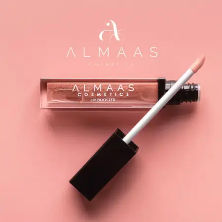 שפופרת של גלוס מנפח שפתיים בגוון ניוד מונחת על משטח ורוד, כשמקל המריחה בעל הכובע השחור מונח באלכסון. לוגו ALMAAS Cosmetics מופיע מעל המוצר באותיות לבנות. | אלמאס קוסמטיקה – מתנה מושלמת של טיפוח ויוקרה