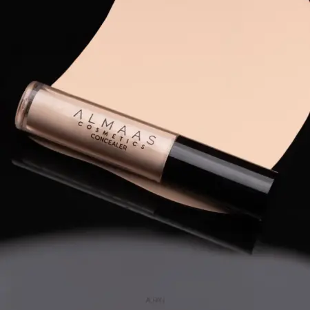 שפופרת של קונסילר בעל כיסוי מלא ומרקם קליל - CONCEALER HD עם מכסה שחור מונחת באלכסון על משטח שחור ומלוטש, כשמאחוריה דוגמית של קונסילר בצבע בז' המנוגדת לרקע המבריק. תווית המוצר נראית בבירור. | אלמאס קוסמטיקה – מתנה מושלמת של טיפוח ויוקרה