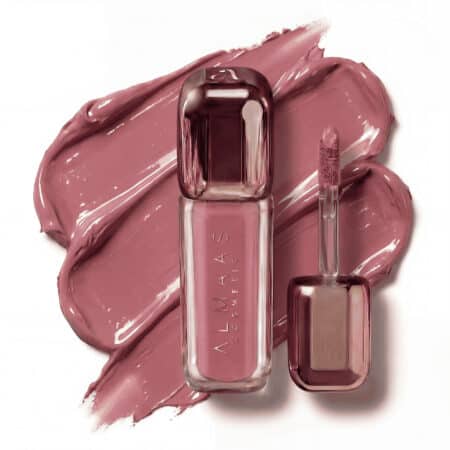 בקבוק שפתון נוזלי בגוון ורוד-סגול הנושא את הכיתוב "ALMAAS Cosmetics" מונח לצד מברשת מריחה תואמת. כתמים של אותו גוון שפתון סגול מכסים את הרקע הלבן, ויוצרים מראה מסוגנן והרמוני. | אלמאס קוסמטיקה – מתנה מושלמת של טיפוח ויוקרה