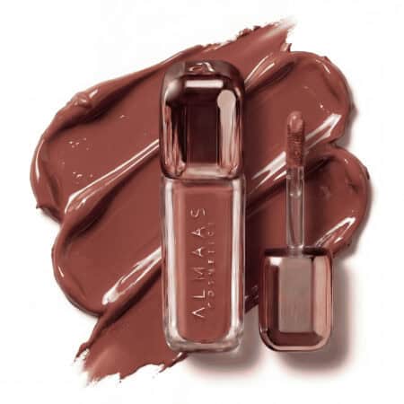 שפופרת בצבע זהב ורוד של שפתון נוזלי מבית ALMAAS Cosmetics, כשמברשת המריחה מונחת לצדה. השפתון נמרח במשיכות רחבות וחלקות על משטח לבן, ומציג גוון חום עשיר וחם. | אלמאס קוסמטיקה – מתנה מושלמת של טיפוח ויוקרה