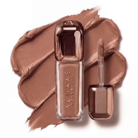 שפתון נוזלי בצבע זהב-ורוד מטאלי, שעליו הכיתוב "ALMAAS COSMETICS", מונח על רקע של שפתון חום-ניוד קרמי ומרוח. מברשת המריחה מונחת לצד השפופרת, ובצבע זהה. המראה אלגנטי ומודרני. | אלמאס קוסמטיקה – מתנה מושלמת של טיפוח ויוקרה