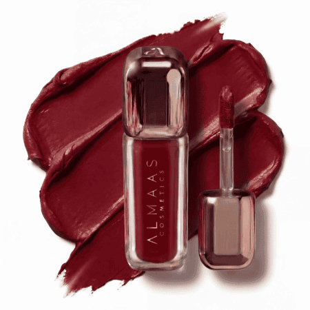 בתמונה נראית שפופרת של שפתון נוזלי מבית ALMAAS Cosmetics בתוך אריזה שקופה בגוון אדום כהה. מברשת המריחה מונחת לצד השפופרת. שתיהן מונחות על כתם באותו גוון אדום עמוק של השפתון, על רקע לבן. | אלמאס קוסמטיקה – מתנה מושלמת של טיפוח ויוקרה