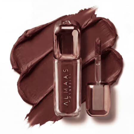 שפופרת אלגנטית בצבע זהב ורוד של שפתון נוזלי, שעליה הכיתוב "ALMAAS COSMETICS", מונחת על כתם של שפתון חום מט. מכסה השפופרת פתוח, ומקל המריחה שעליו נותר חומר חום מונח לצדה על רקע מחוספס. | אלמאס קוסמטיקה – מתנה מושלמת של טיפוח ויוקרה
