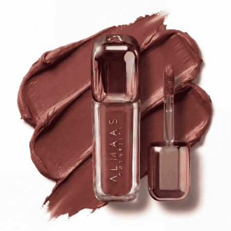 שפופרת שקופה של שפתון נוזלי מבית ALMAAS COSMETICS, המצוידת במברשת מריחה בצורת כפה, מונחת על כתמי שפתון בגוון חום-סגול. השפופרת והמברשת מסודרות באלכסון על רקע לבן, ומדגישות את המרקם הקרמי והצבע העשיר. | אלמאס קוסמטיקה – מתנה מושלמת של טיפוח ויוקרה