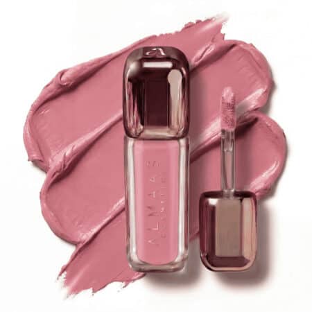 שפתון נוזלי בגוון ורוד-סגול, בתוך שפופרת מלבנית שקופה עם מכסה מוזהב-ורוד מבריק, מונח על כתם שפתון תואם. מברשת המריחה, המונחת לצד השפופרת, בעלת קצה רך ומזווית המצופה באותו צבע. על התווית כתוב "ALMAAS COSMETICS". | אלמאס קוסמטיקה – מתנה מושלמת של טיפוח ויוקרה