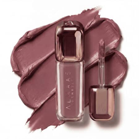 בקבוק שפתון נוזלי בגוון ורוד-סגול, שעליו הכיתוב "ALMAAS COSMETICS", מונח על כתם שפתון באותו הגוון. מברשת המריחה, המכוסה באותו הגוון, מונחת לצד הבקבוק. האריזה מעוצבת בקווים נקיים, עם אלמנטים מתכתיים ושקופים. | אלמאס קוסמטיקה – מתנה מושלמת של טיפוח ויוקרה