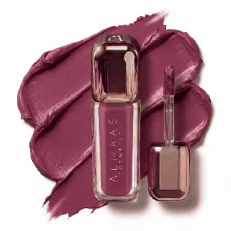 שפתון נוזלי בגוון ורוד-סגול, בתוך שפופרת אלגנטית ומבריקה שעליה הכיתוב "ALMAAS COSMETICS", מונח על כתם בצבע תואם. המברשת, המכוסה באותו הגוון, מונחת לצד השפופרת על רקע קרמי ומרקם. | אלמאס קוסמטיקה – מתנה מושלמת של טיפוח ויוקרה