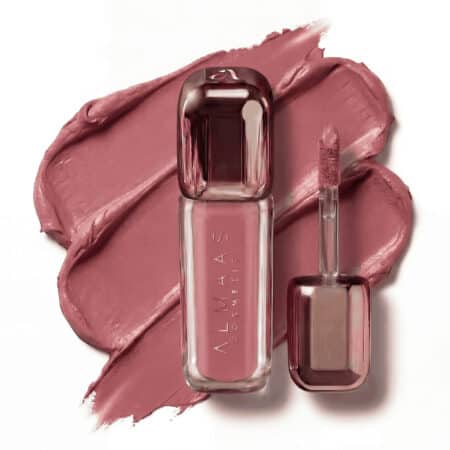 שפופרת זכוכית מלוטשת המכילה שפתון נוזלי בגוון סגול-ורוד, שעליה כתוב "Almaas Cosmetics", ניצבת לצד מברשת המריחה שלה, ושניהם מונחים על כתם מרקם באותו גוון סגול-ורוד. המראה הכללי מבריק ואלגנטי, ומדגיש את הצבע העשיר של השפתון. | אלמאס קוסמטיקה – מתנה מושלמת של טיפוח ויוקרה