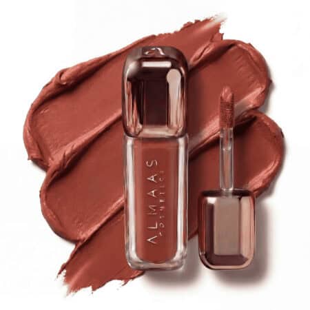 שפופרת גלוס לשפתיים בצבע זהב ורוד מטאלי, שעליה הכיתוב "ALMAAS COSMETICS", מונחת על גבי כתם של שפתון טרקוטה עשיר ומט. לצדה, מוצג המברשת התואמת עם חומר המוצר על קצהו. הרקע לבן. | אלמאס קוסמטיקה – מתנה מושלמת של טיפוח ויוקרה
