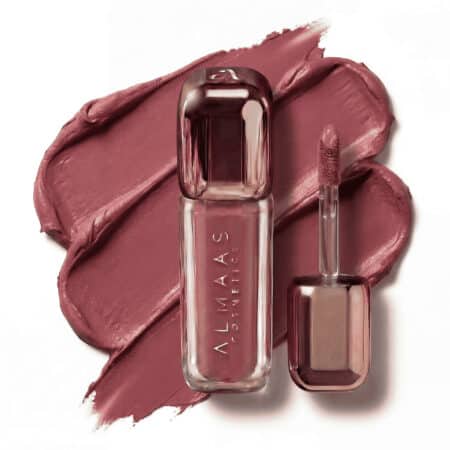 שפתון נוזלי בגוון סגול-ורוד מבית ALMAAS Cosmetics מונח על כתם סגול-ורוד תואם. הבקבוק המלוטש בגוון ורדרד מונח לצד מברשת המריחה, שבראשה קצה ספוג רך המצופה בשפתון. המראה הכללי הוא מודרני ואלגנטי. | אלמאס קוסמטיקה – מתנה מושלמת של טיפוח ויוקרה