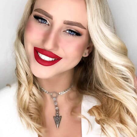 אישה עם שיער בלונדיני ארוך וגלי מחייכת למצלמה. היא עוטה שפתון אדום בוהק, איפור עיניים דרמטי ושרשרת בולטת עם תליון משולש. היא לבושה בבגדים לבנים, והרקע הוא בצבע בהיר ונטול דוגמאות. | אלמאס קוסמטיקה – מתנה מושלמת של טיפוח ויוקרה