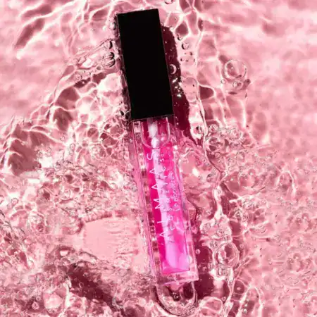 שפופרת גלוס שקופה עם מכסה שחור מונחת על משטח ורוד, דליל ומלא בועות. הנוזל הוורוד שבתוך השפופרת משתלב עם הרקע הוורוד העז והמבריק, ויוצר מראה נוצץ ויוקרתי. | אלמאס קוסמטיקה – מתנה מושלמת של טיפוח ויוקרה