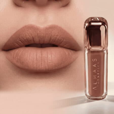 תקריב של שפתי אישה המורחת שפתון ניוד מט. לצד השפתיים מונח שפופרת שקופה של שפתון מבית Almas Cosmetics בגוון ניוד תואם, עם לוגו לבן על הבקבוק. הרקע הוא בגוון בז' רך. | אלמאס קוסמטיקה – מתנה מושלמת של טיפוח ויוקרה