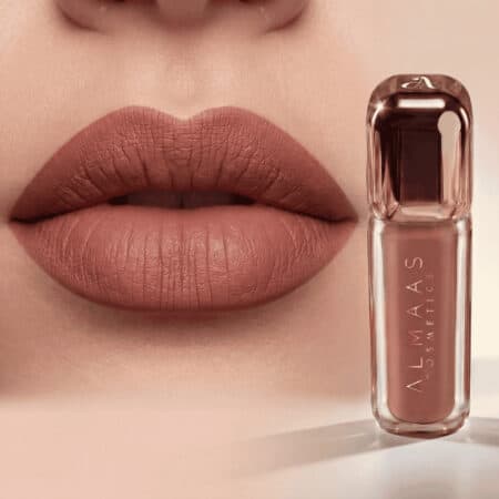 תקריב של שפתי אישה המורחות בשפתון ניוד מט, כשהן פתוחות קלות. לצד השפתיים מונח שפופרת שקופה של שפתון מבית ALMAAS Cosmetics בגוון ניוד תואם, באריזה אלגנטית ומודרנית עם לוגו לבן על הבקבוק. רקע בהיר. | אלמאס קוסמטיקה – מתנה מושלמת של טיפוח ויוקרה