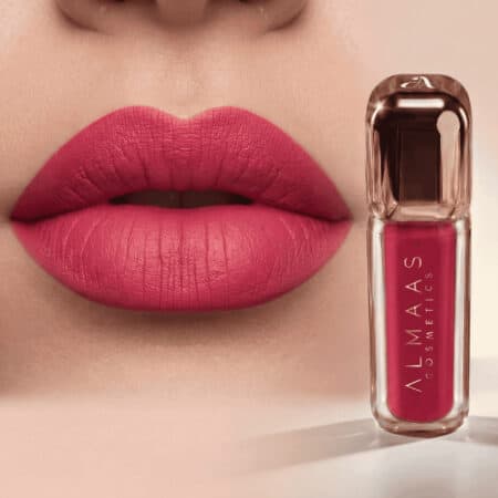 תקריב של שפתיים המרוחות בשפתון ורוד עז במראה מט. לצד השפתיים מונחת שפופרת שפתון ורודה תואמת, שעליה כתוב "ALMAAS COSMETICS" באותיות לבנות, על רקע בז' רך ומטושטש. התמונה מדגישה את גוון השפתון ואת אריזתו. | אלמאס קוסמטיקה – מתנה מושלמת של טיפוח ויוקרה