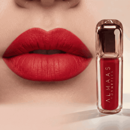 תקריב של שפתי אישה המורחות שפתון אדום מט. לצד פניה, שפופרת שקופה של שפתון נוזלי אדום עם הכיתוב "ALMAAS COSMETICS" ניצבת זקופה על משטח חלק ובהיר. המראה הכללי נועז ואלגנטי. | אלמאס קוסמטיקה – מתנה מושלמת של טיפוח ויוקרה