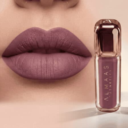 תקריב של שפתיים המרוחות בשפתון סגול-ורוד מט. לצד השפתיים מונחת שפופרת שפתון בצבע סגול-ורוד, ועליה הכיתוב "ALMAAS COSMETICS" באותיות לבנות אנכיות על גבי האריזה השקופה. הרקע רך וניטרלי. | אלמאס קוסמטיקה – מתנה מושלמת של טיפוח ויוקרה