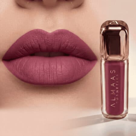 תקריב של שפתי אישה המורחות שפתון סגלגל מט. לצד השפתיים מונח שפופרת שקופה של שפתון נוזלי מבית ALMAAS Cosmetics בגוון סגלגל תואם, עם מכסה זהוב ושם המותג מודפס אנכית על הבקבוק. | אלמאס קוסמטיקה – מתנה מושלמת של טיפוח ויוקרה