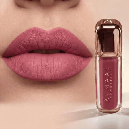 תקריב של שפתי אישה המורחות בשפתון ורוד מט. לצד השפתיים מונחת שפופרת של שפתון נוזלי מבית ALMAAS Cosmetics בגוון ורוד תואם, בבקבוק מלבני שקוף עם מכסה בצבע זהב ורוד, המוצגת על משטח בהיר. | אלמאס קוסמטיקה – מתנה מושלמת של טיפוח ויוקרה